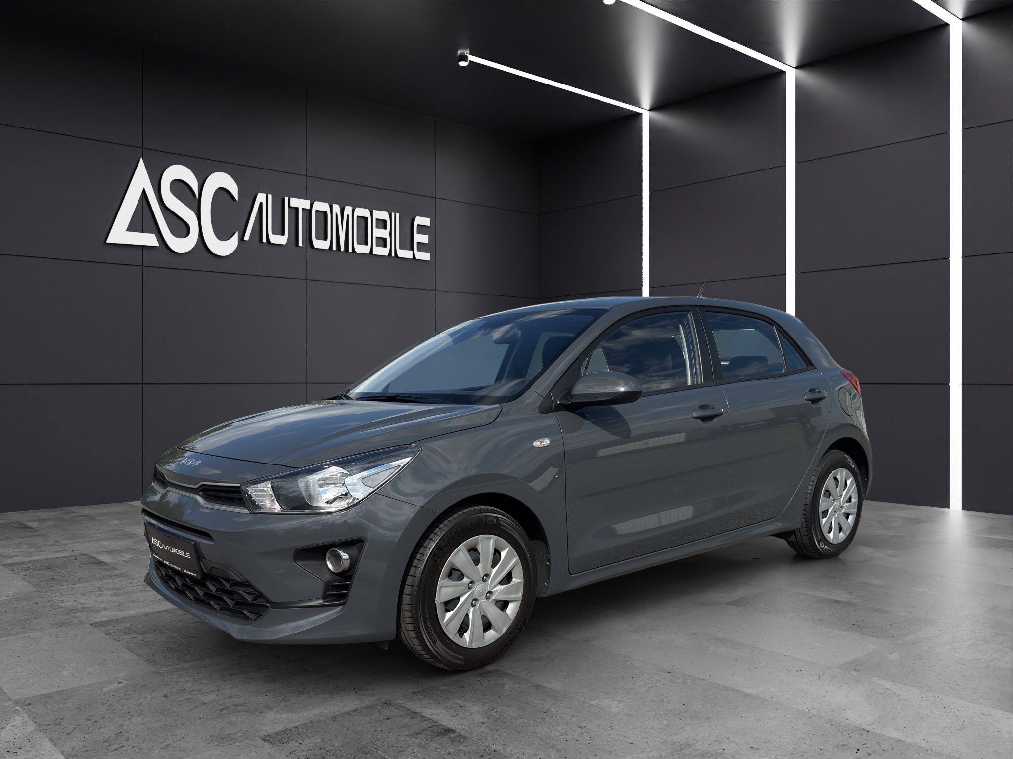 KIA Rio in Dunkelgrau, schräg von vorne links im Showroom, Fünftürer mit silbernen Radkappen